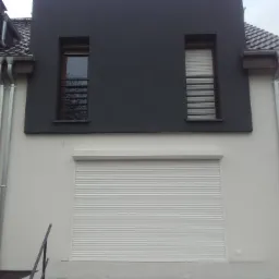 Biała brama rolowana, zamknięta, w nowoczesnym domu z szarą elewacją i metalowymi rynnami, widok z dołu.