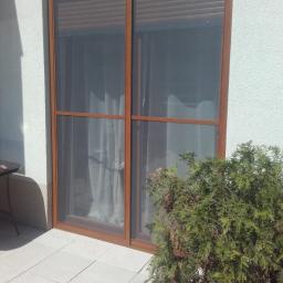 M-PAT Monika Paterska - Brązowe okno z zasłoniętą roletą, widok z zewnątrz na jasny taras z rośliną iglastą.