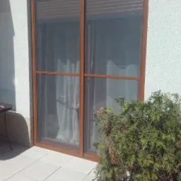 Brązowe okno z zasłoniętą roletą, widok z zewnątrz na jasny taras z rośliną iglastą.