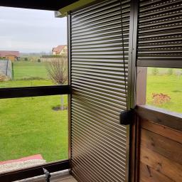 M-PAT Monika Paterska - Montaż brązowej rolety zewnętrznej na tarasie z drewnianą balustradą, widoczne deski i poziomica, krajobraz wiejski w tle.