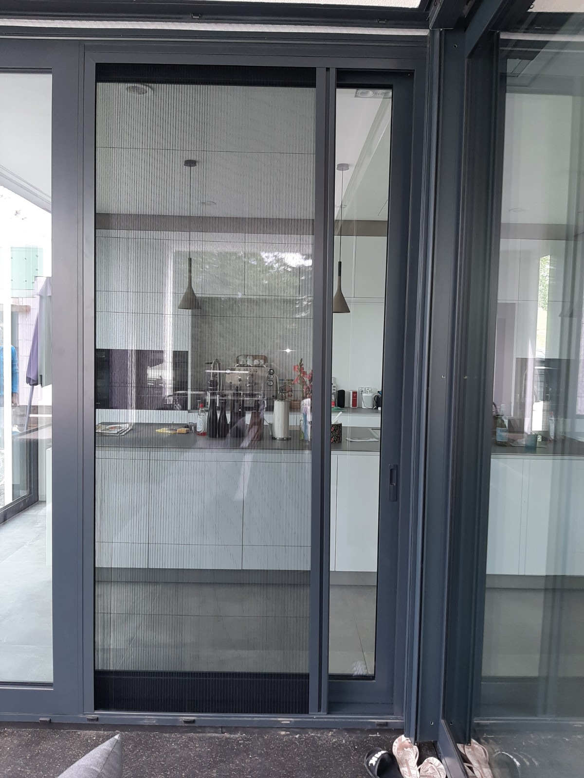 Zasłonięte moskitierą przesuwne drzwi tarasowe z szarą ramą, widok na nowoczesną kuchnię z wiszącymi lampami.