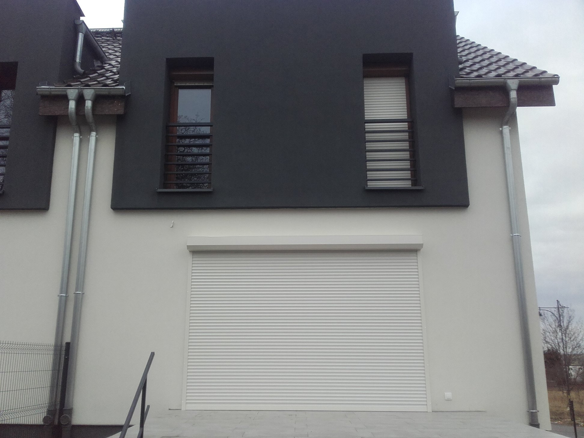 Biała brama rolowana, zamknięta, w nowoczesnym domu z szarą elewacją i metalowymi rynnami, widok z dołu.