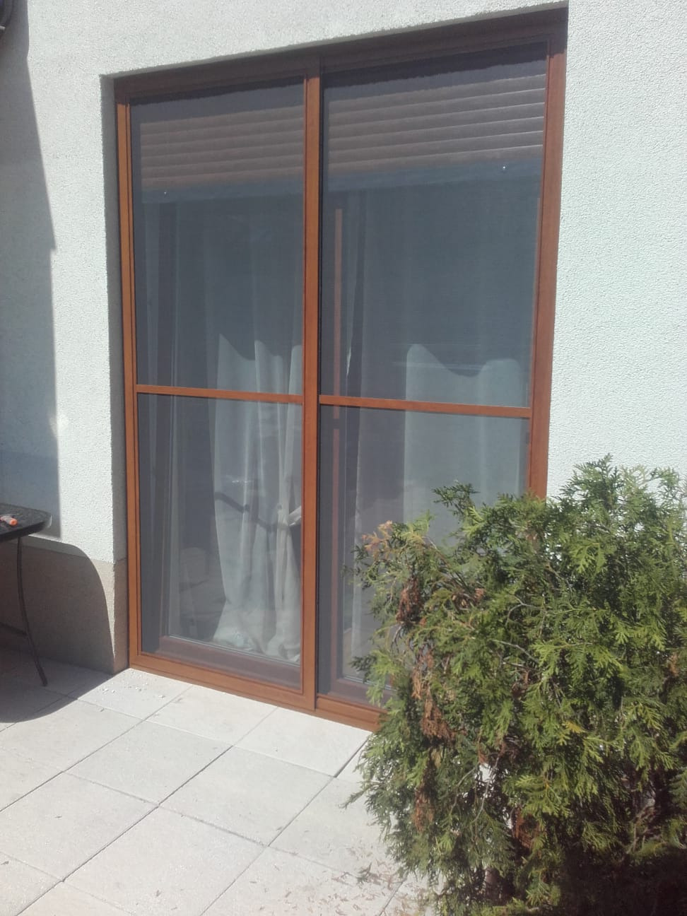 Brązowe okno z zasłoniętą roletą, widok z zewnątrz na jasny taras z rośliną iglastą.