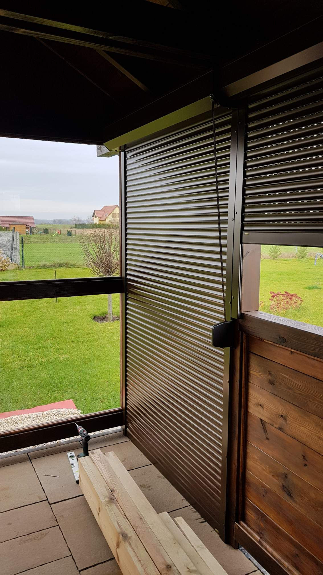 Montaż brązowej rolety zewnętrznej na tarasie z drewnianą balustradą, widoczne deski i poziomica, krajobraz wiejski w tle.