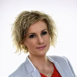 Portret kobiety z kręconymi blond włosami, w marynarce w paski i czerwonej bluzce, z delikatną biżuterią na białym tle.