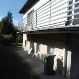 Dwupiętrowy dom z szarym balkonem i ciemnymi rynnami, widoczny z boku, z zielonym koszem na śmieci na pierwszym planie.