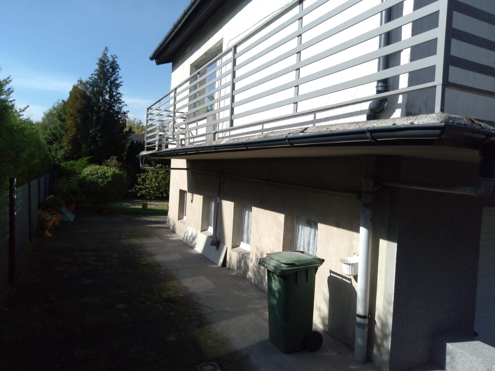 Dwupiętrowy dom z szarym balkonem i ciemnymi rynnami, widoczny z boku, z zielonym koszem na śmieci na pierwszym planie.