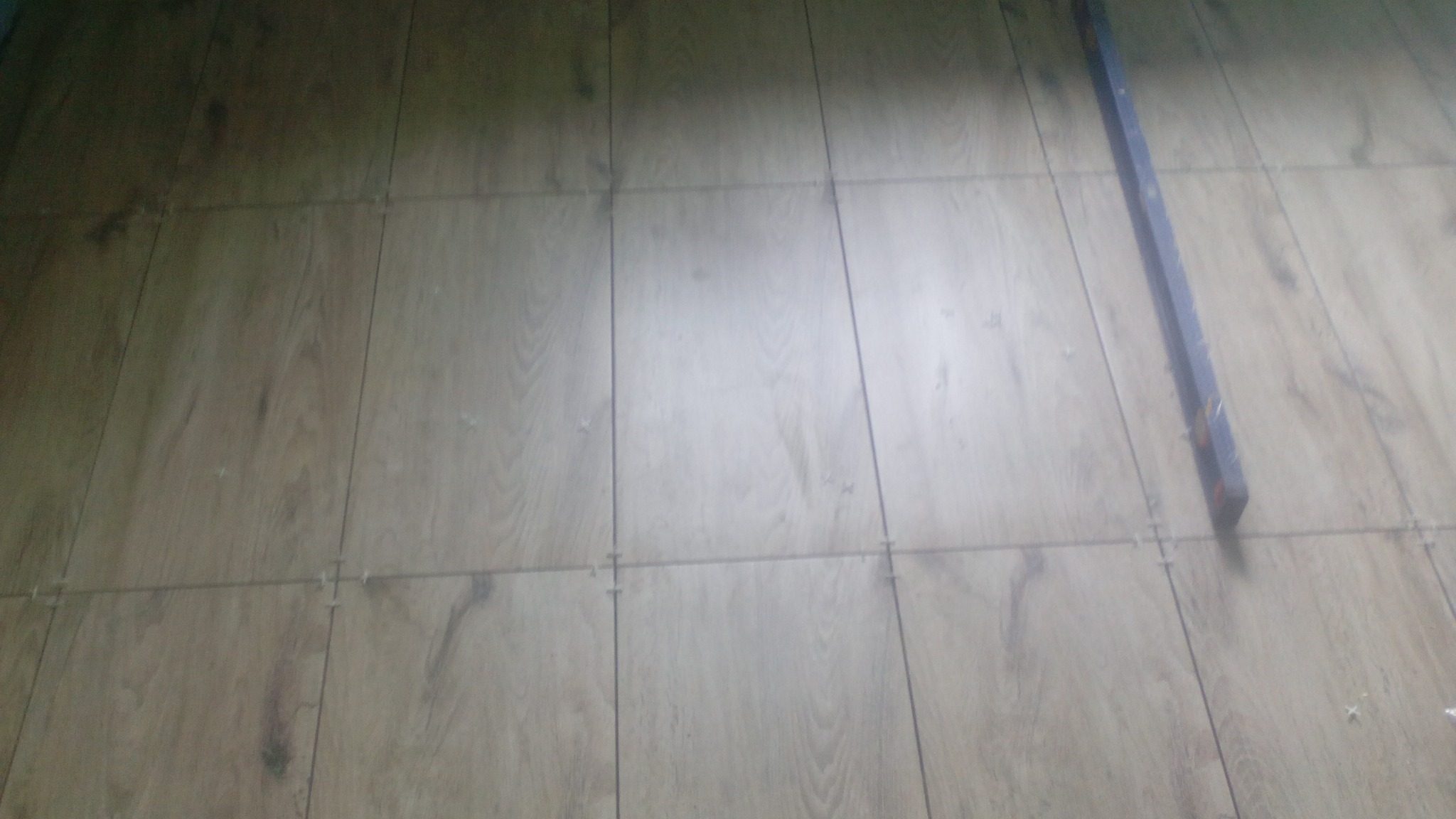 Układanie paneli podłogowych z widocznymi krzyżykami dystansowymi i poziomicą, w trakcie montażu, słabe oświetlenie.