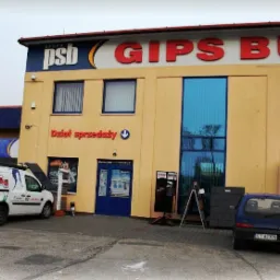 Fasada budynku składu budowlanego Grupa PSB GIPS BUD z napisem 'Dział sprzedaży', zaparkowany biały bus z reklamami farb i ociepleń oraz niebieski samochód osobowy.