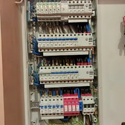 Rozdzielnia elektryczna z zainstalowanymi bezpiecznikami, wyłącznikami różnicowoprądowymi i ogranicznikami przepięć, widoczne przewody w kolorach niebieskim, czarnym, brązowym i żółto-zielonym...