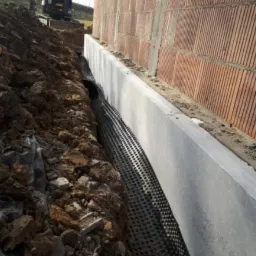 Fundament budynku w trakcie budowy z wykonanym zabezpieczeniem i drenażem; widoczna perforowana rura drenażowa w wykopie, zabezpieczona folią fundamentowa, w tle budynek z cegły i koparka.