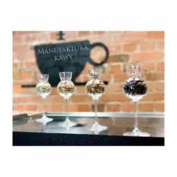 Cztery kieliszki na wysokich nóżkach wypełnione różnymi rodzajami ziaren kawy, ustawione na czarnym blacie, w tle logo Manufaktury Kawy na ceglanej ścianie.
