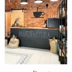 Wnętrze kawiarni z ceglaną ścianą, czarnym blatem z napisem 'MANUFAKTURA', workami z kawą i trzema lampami wiszącymi. Dekoracyjna czarna filiżanka na ścianie.