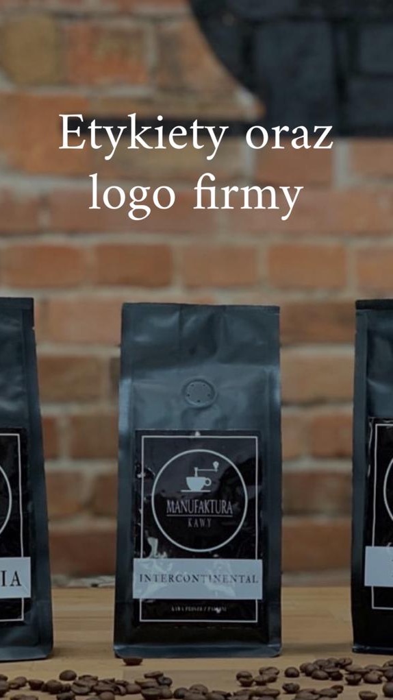 Trzy czarne opakowania kawy z logo 'Manufaktura Kawy Intercontinental' na drewnianym blacie z rozsypanymi ziarnami kawy, na tle ściany z czerwonej cegły.