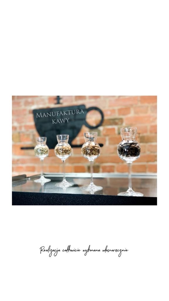 Cztery kieliszki na wysokich nóżkach wypełnione różnymi rodzajami ziaren kawy, ustawione na czarnym blacie, w tle logo Manufaktury Kawy na ceglanej ścianie.