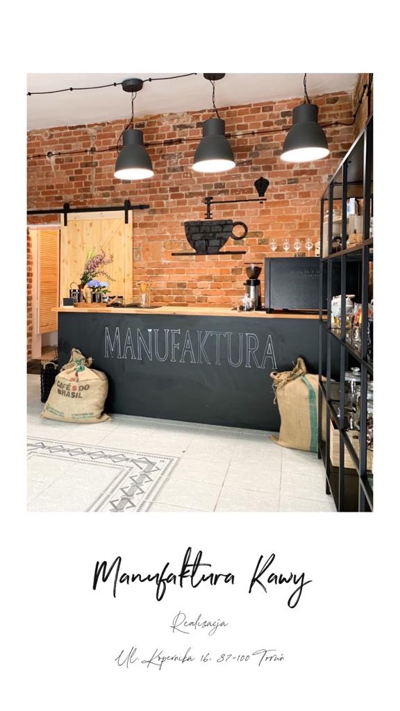 Wnętrze kawiarni z ceglaną ścianą, czarnym blatem z napisem 'MANUFAKTURA', workami z kawą i trzema lampami wiszącymi. Dekoracyjna czarna filiżanka na ścianie.