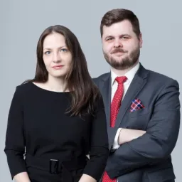 Portret dwojga prawników: kobieta w czarnej sukience z paskiem i mężczyzna w szarym garniturze z czerwoną koszulą i wzorzystą poszetką, na jednolitym szarym tle.