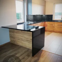 Nowoczesna kuchnia z wyspą o blacie z czarnego kamienia i drewnianym wykończeniem, widok z boku, w tle szafki kuchenne z drewnianymi frontami i czarną mozaiką nad blatem.