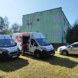 Gedeon Wróblewski Sp.j - Dwa białe busy firmy Gedeon z logo i danymi kontaktowymi zaparkowane na trawie przed budynkiem z czerwoną rurą wentylacyjną prowadzącą do okna na poddaszu, obok stoi osobowy samochód z logo firmy.