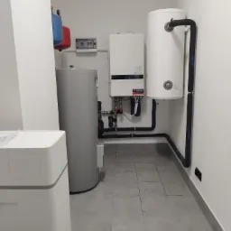Kotłownia z nowoczesnym piecem gazowym, zasobnikiem ciepłej wody, zbiornikiem wyrównawczym i zmiękczaczem wody w minimalistycznym wnętrzu z szarymi płytkami podłogowymi.