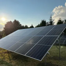 Panel słoneczny zainstalowany na trawiastym terenie, z widocznym słońcem i drzewami w tle, uchwycony w delikatnym świetle popołudniowym.