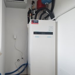 Gedeon Wróblewski Sp.j - Biała pompa ciepła Panasonic Aquarea Smart Cloud zamontowana w pomieszczeniu, z widocznymi niebieskimi rurami, czerwonym i niebieskim zbiornikiem wyrównawczym, oraz szafką elektryczną na ścianie.