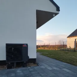 Czarna jednostka zewnętrzna z logo Gedeon zamontowana przy białej ścianie domu, widoczna kostka brukowa i trawnik w tle.