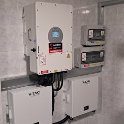 Gedeon Wróblewski Sp.j - Instalacja fotowoltaiczna w pomieszczeniu: falownik Deye z logo Gedeon, dwa moduły V-TAC oraz rozdzielnice elektryczne na ścianie wyłożonej płytkami.