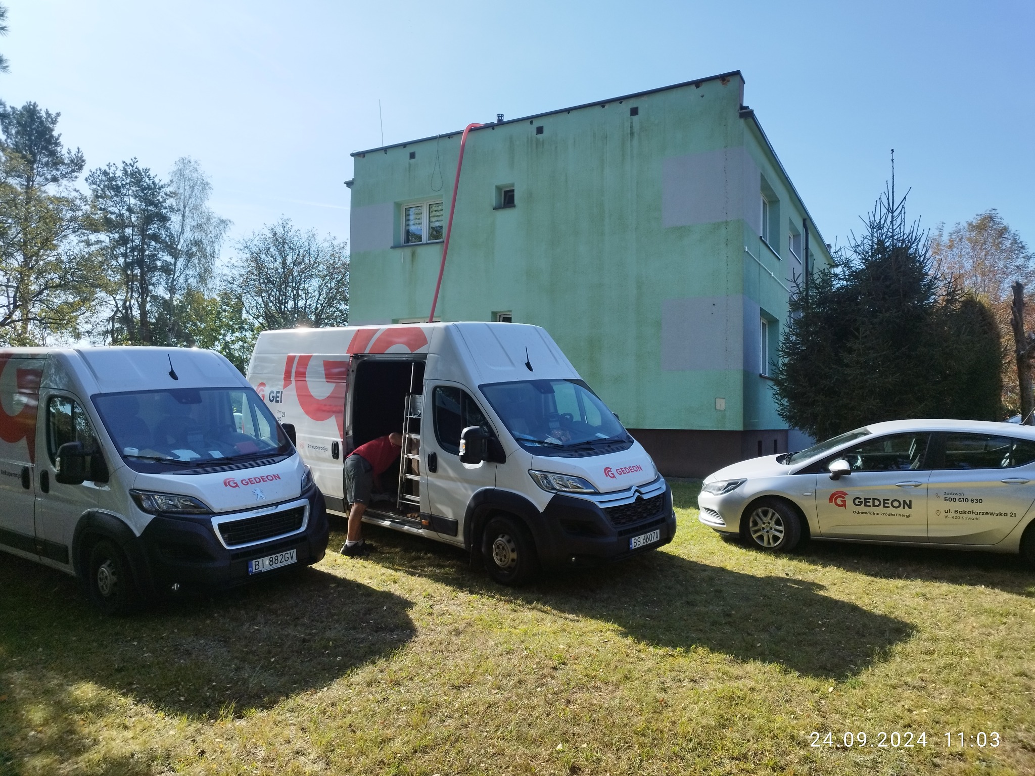 Dwa białe busy firmy Gedeon z logo i danymi kontaktowymi zaparkowane na trawie przed budynkiem z czerwoną rurą wentylacyjną prowadzącą do okna na poddaszu, obok stoi osobowy samochód z logo firmy.