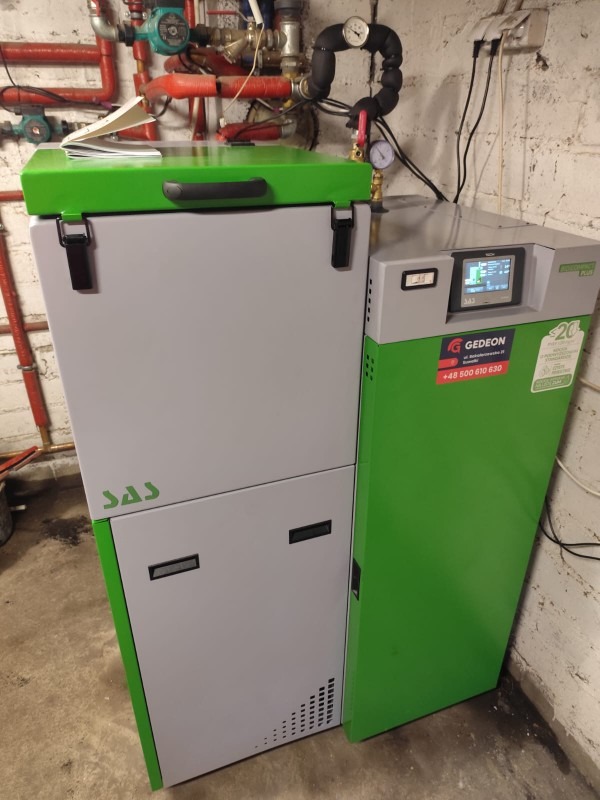 Szaro-zielony piec na paliwo stałe SAS Gedeon PLUS z widocznym panelem sterowania i instalacją hydrauliczną w tle, zainstalowany w piwnicy.
