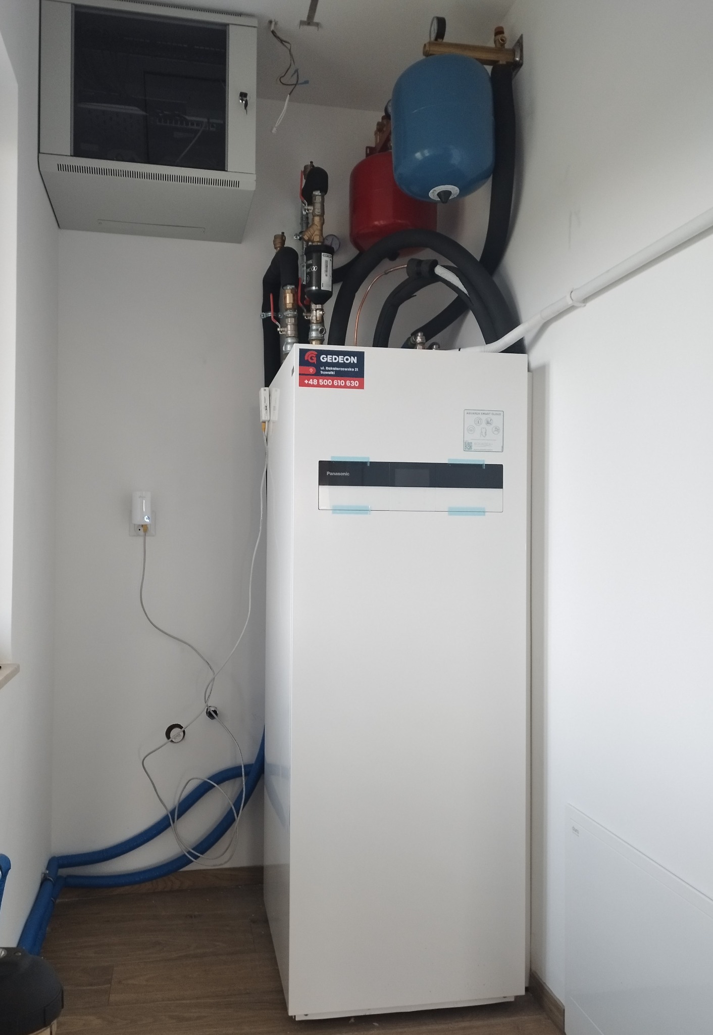 Biała pompa ciepła Panasonic Aquarea Smart Cloud zamontowana w pomieszczeniu, z widocznymi niebieskimi rurami, czerwonym i niebieskim zbiornikiem wyrównawczym, oraz szafką elektryczną na ścianie.