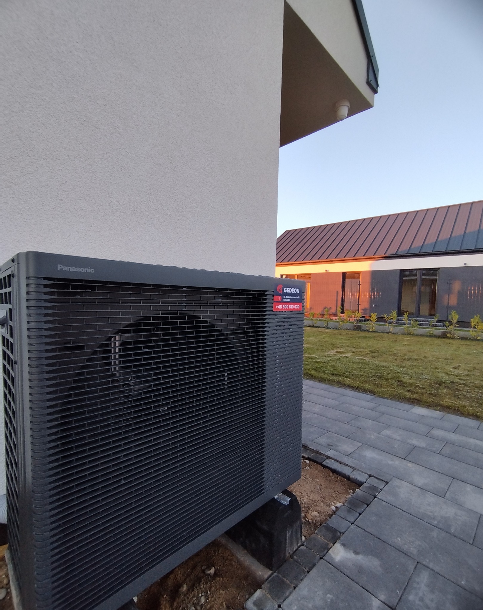 Czarna jednostka zewnętrzna klimatyzacji Panasonic zamontowana przy ścianie domu z kostki brukowej. Na urządzeniu naklejka z logo i numerem telefonu firmy Gedeon.
