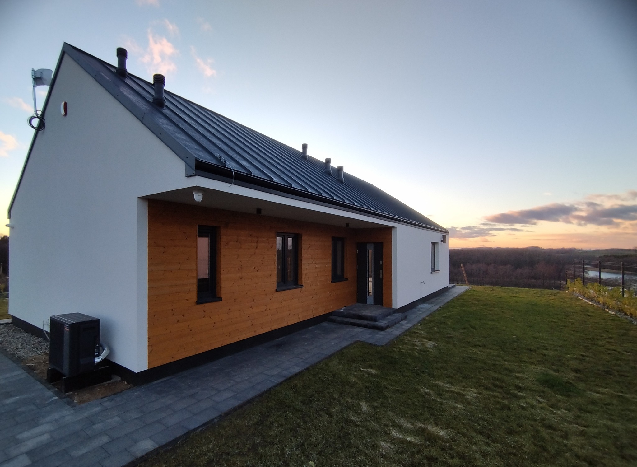 Nowoczesny dom jednorodzinny z drewnianą elewacją, czarnym dachem z blachy trapezowej i zamontowaną pompą ciepła na zewnątrz, widok na zachodzące słońce w tle.