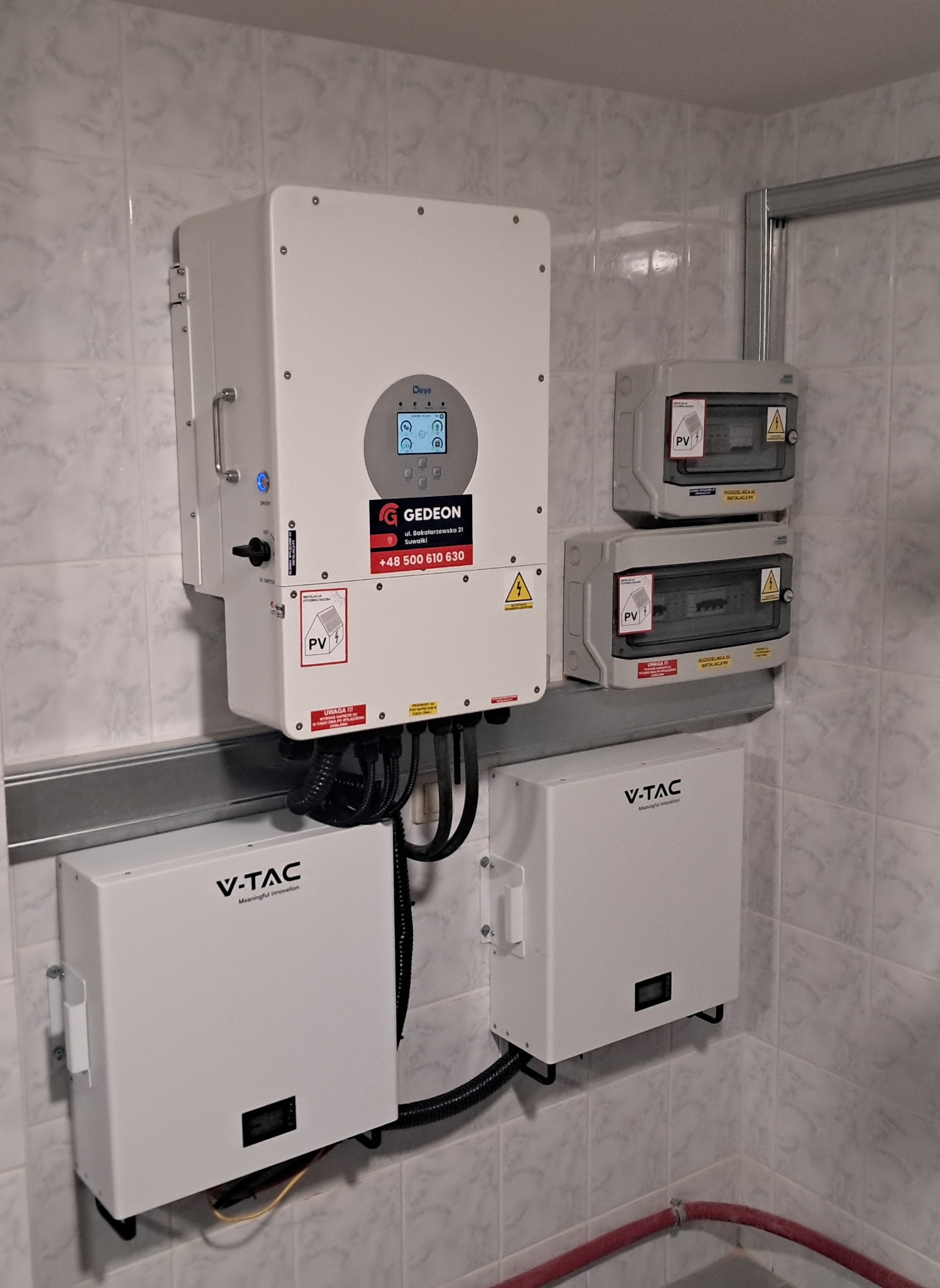 Instalacja fotowoltaiczna w pomieszczeniu: falownik Deye z logo Gedeon, dwa moduły V-TAC oraz rozdzielnice elektryczne na ścianie wyłożonej płytkami.