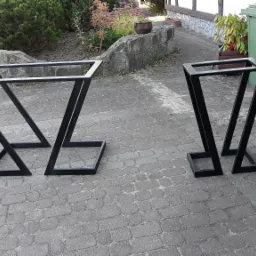 Dwie czarne, metalowe konstrukcje o geometrycznym kształcie, prawdopodobnie podstawy stołów, stoją na szarej kostce brukowej na zewnątrz budynku.