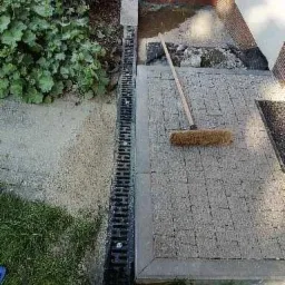 Fragment posesji z ułożoną szarą kostką brukową, widoczny odpływ liniowy, częściowo ułożona kostka i leżąca zmiotka.