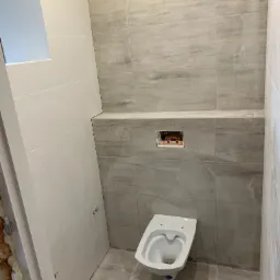 Zabudowa toalety podtynkowej z szarymi płytkami imitującymi drewno, biała miska wisząca, białe płytki na ścianie obok drzwi z pianką montażową w ościeżnicy.