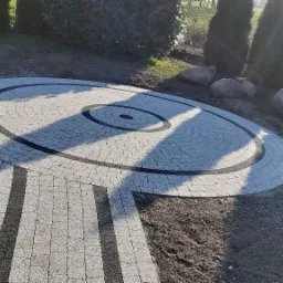 Okrągła powierzchnia z kostki brukowej w ogrodzie, z ciemnymi obwódkami i ścieżką z kostki prowadzącą do niej, otoczona zielenią i kamieniami, w słoneczny dzień.