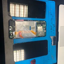 Rozłożony smartfon z uszkodzonym ekranem, widoczna płyta gł&oacute;wna, odłączony moduł aparatu, narzędzia i śrubki na niebieskiej macie serwisowej.