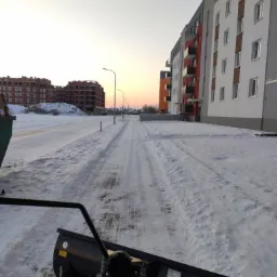 Widok z perspektywy operatora na pług śnieżny podczas odśnieżania drogi osiedlowej, w tle bloki mieszkalne i budowa w zimowej scenerii o zachodzie słońca.