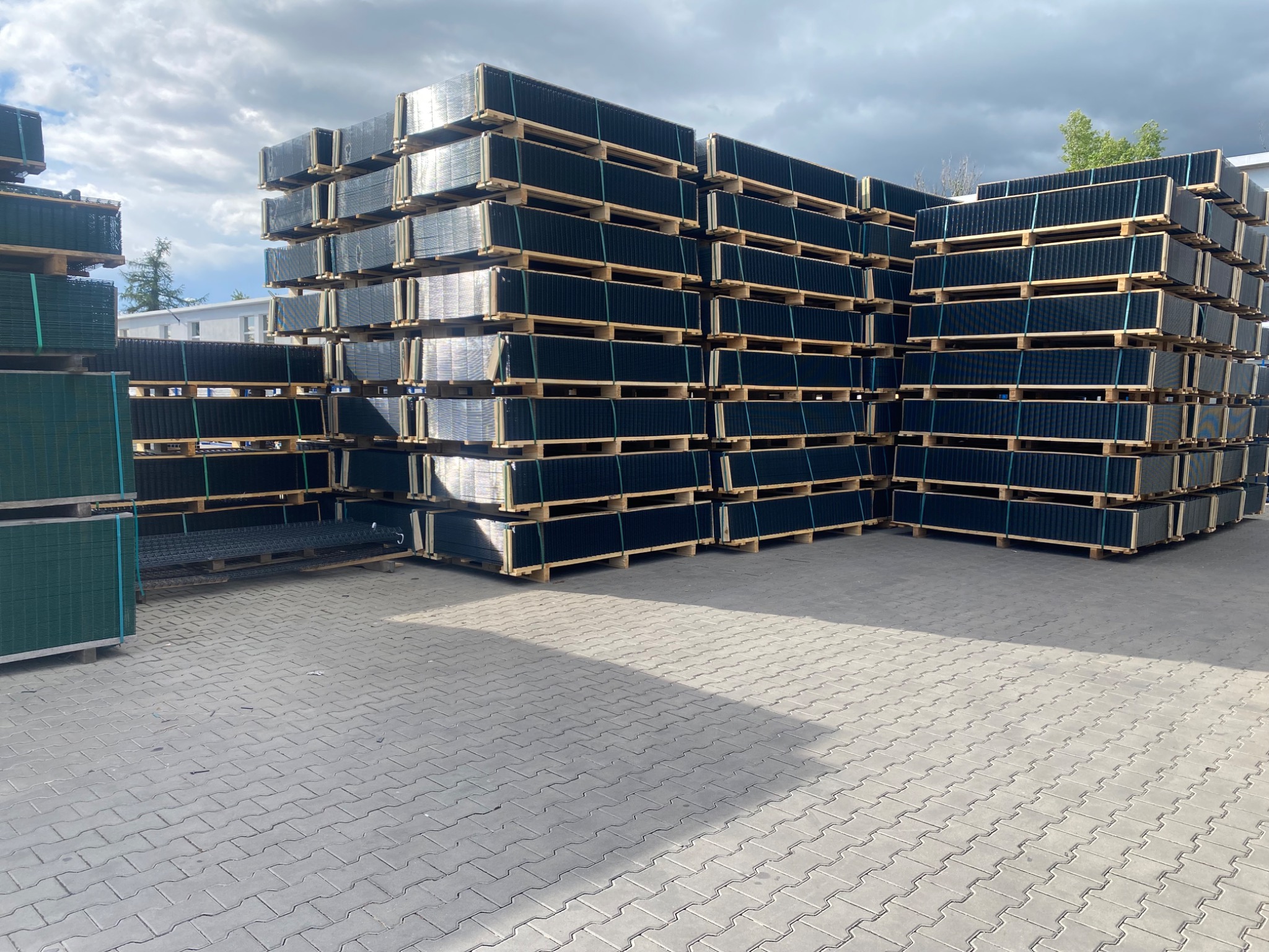 Stosy paneli ogrodzeniowych, zabezpieczonych taśmami, ułożone na paletach na kostce brukowej. Widoczne budynki w tle i zachmurzone niebo.
