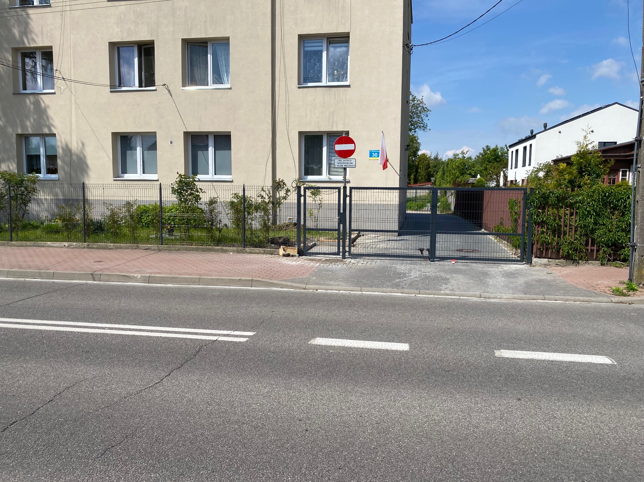 Szara brama wjazdowa i ogrodzenie panelowe przed budynkiem z tabliczką 'zakaz wjazdu' i polską flagą. Widok z ulicy.