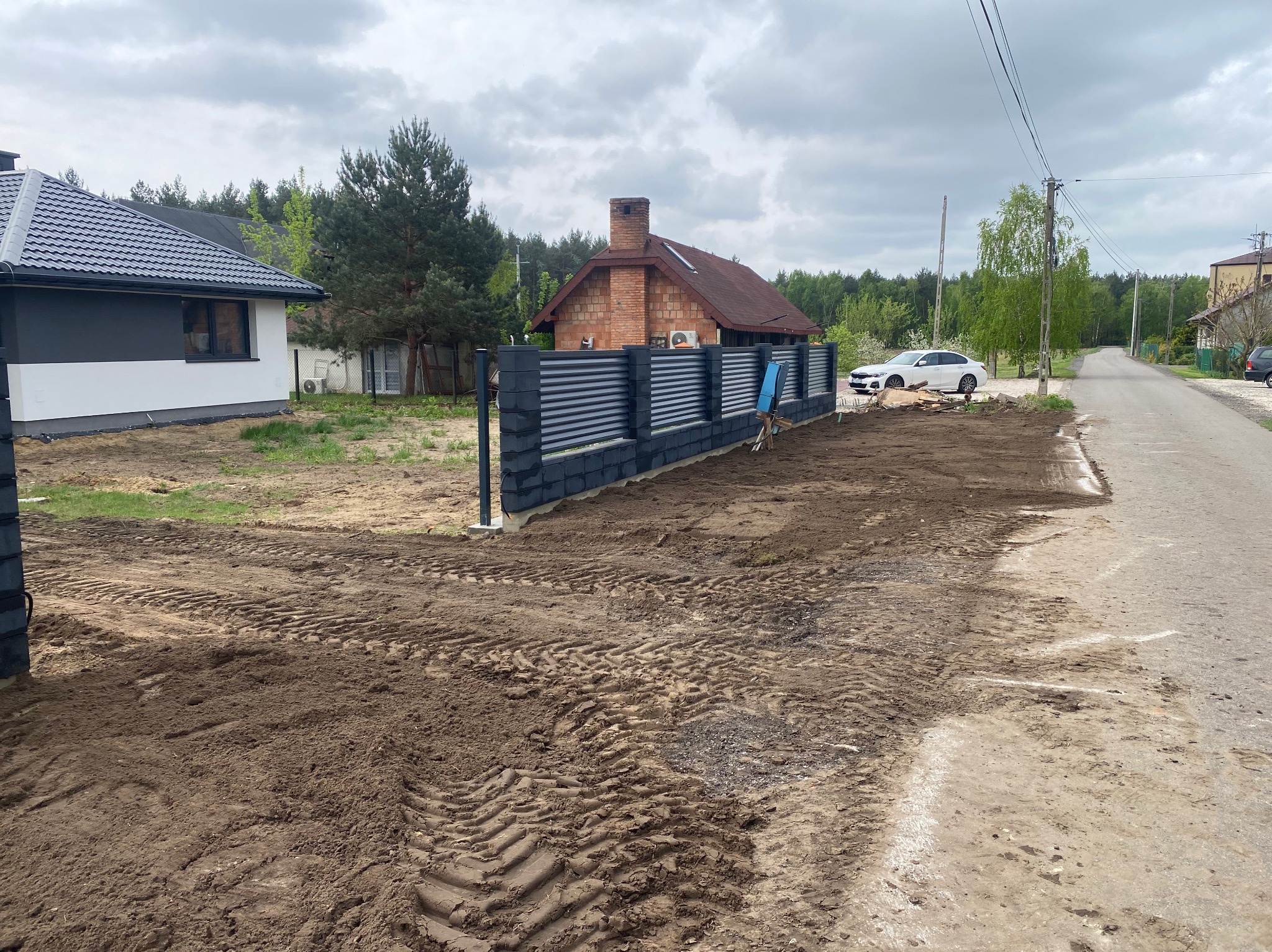 Nowoczesne ogrodzenie panelowe z grafitowymi słupkami i poziomymi przęsłami, widoczne ślady opon na ziemi po montażu, dom w tle. Ujęcie z zewnątrz.