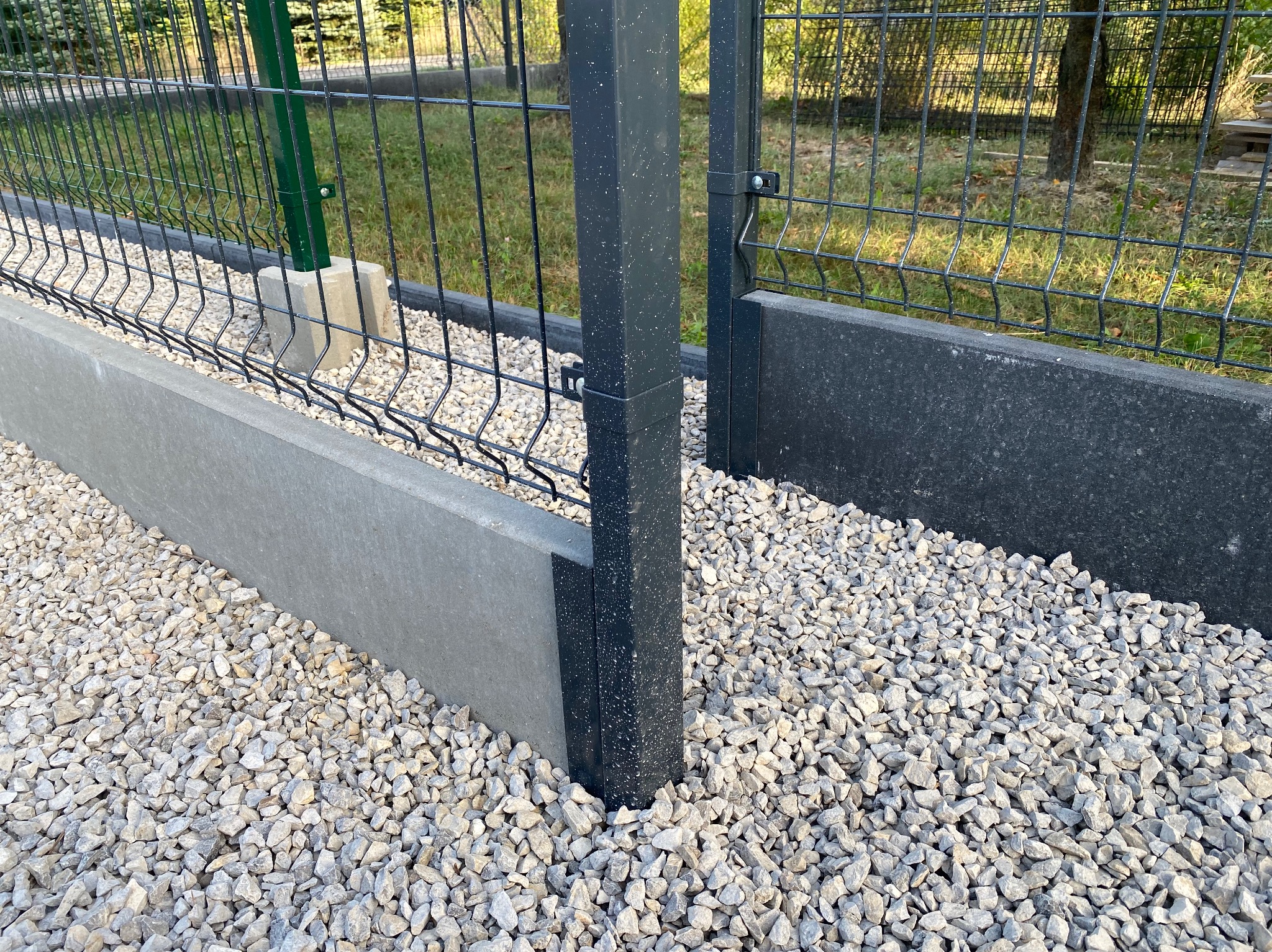 Fragment zamontowanego, panelowego ogrodzenia z podmurówką i słupkami. Białe kamienie dekoracyjne u podstawy. Widok z bliska na gotowy fragment ogrodzenia.