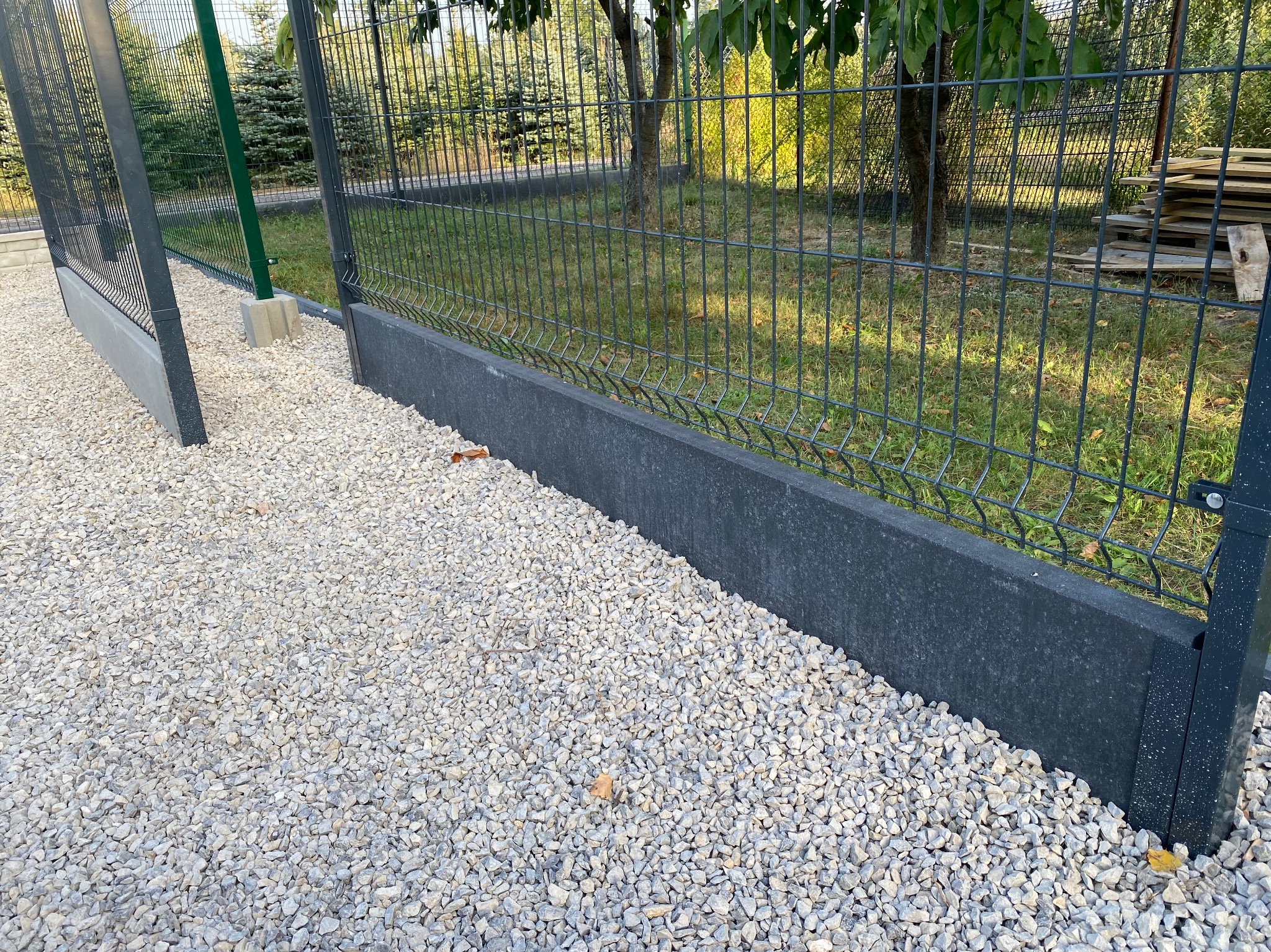 Fragment panelowego ogrodzenia z podmurówką, wysypane kamieniami, widok z bliska. Elementy metalowe w kolorze grafitowym, słupki zielone.