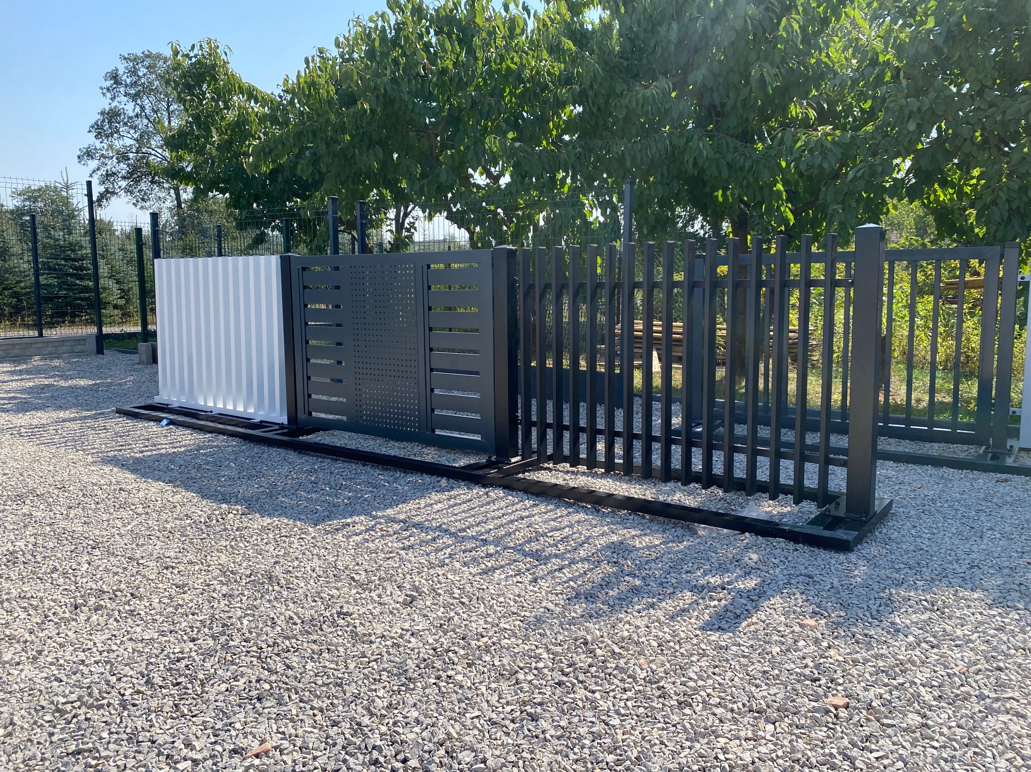 Nowoczesna brama przesuwna z metalowymi panelami w kolorze grafitowym i białym, na żwirowym podłożu, w tle zieleń drzew. Minimalistyczny design.