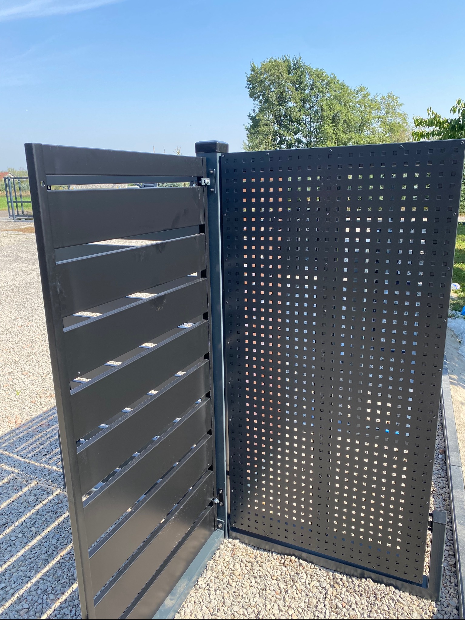 Nowoczesna, otwarta brama wjazdowa z czarnego metalu z perforowanym panelem i poziomymi szczelinami. Widoczny mechanizm zawiasów i otoczenie z kamyków.