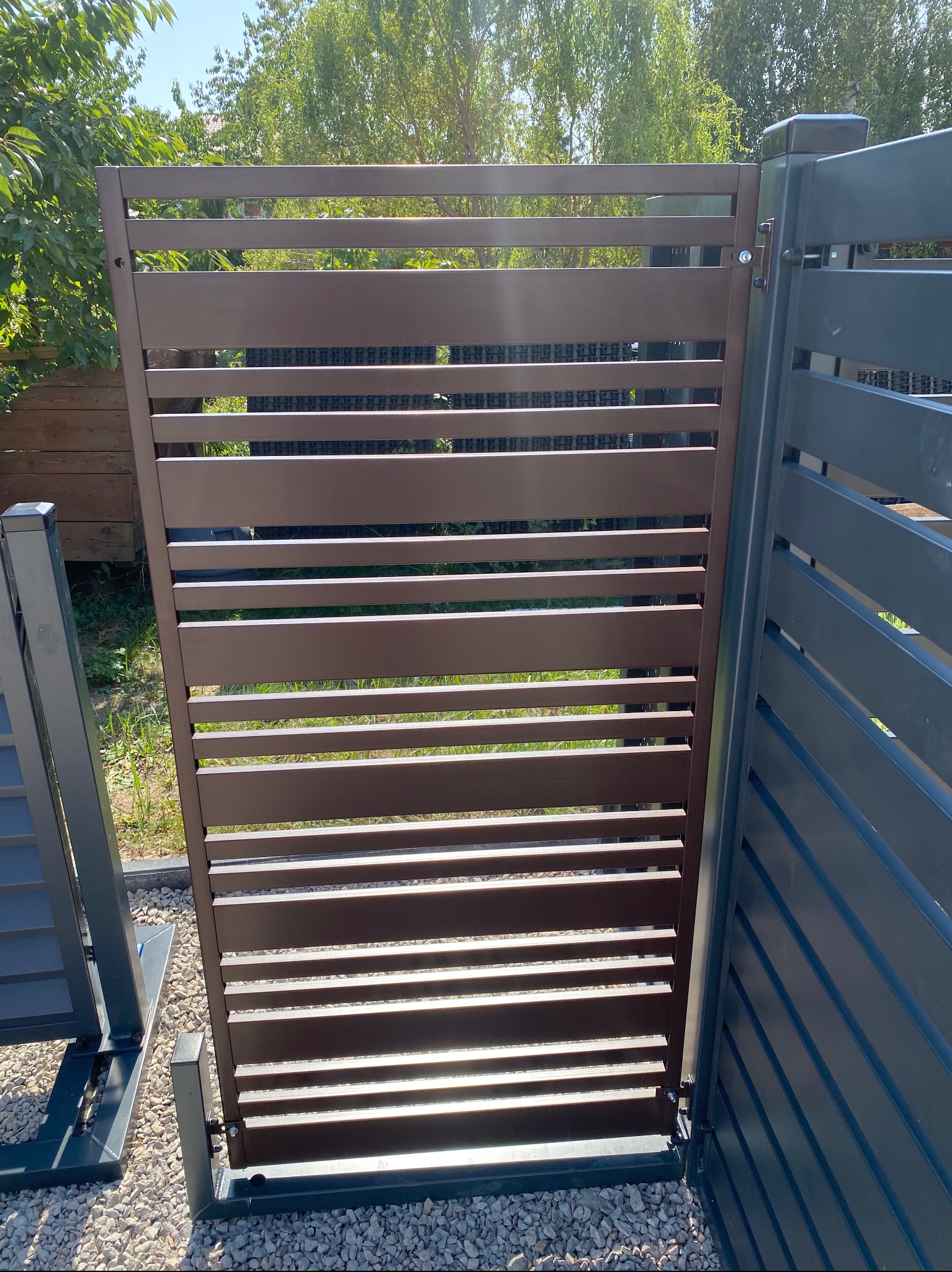 Nowoczesne, poziome ogrodzenie panelowe w kolorze brązowym, zamontowane na posesji z zielenią w tle. Widoczne detale montażu i fragment sąsiedniego, szarego panelu.