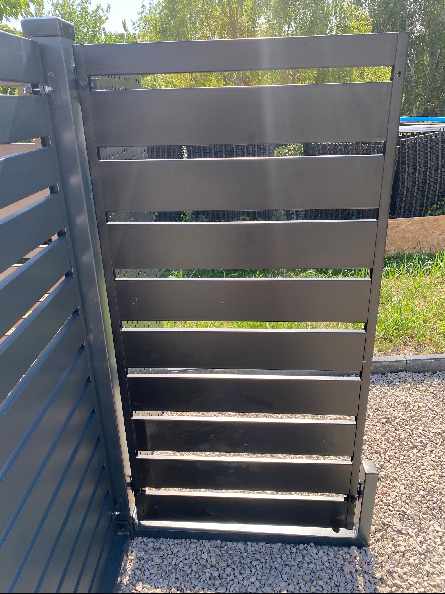 Nowoczesna, grafitowa furtka z poziomych paneli metalowych, widoczna z bliska na tle zieleni i jasnego żwiru.