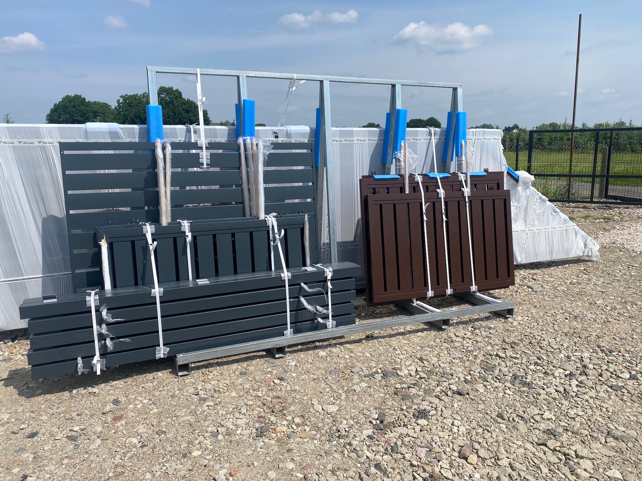 Przygotowane elementy ogrodzenia panelowego: grafitowe i brązowe przęsła na palecie, zabezpieczone folią, gotowe do montażu na żwirowym podłożu.